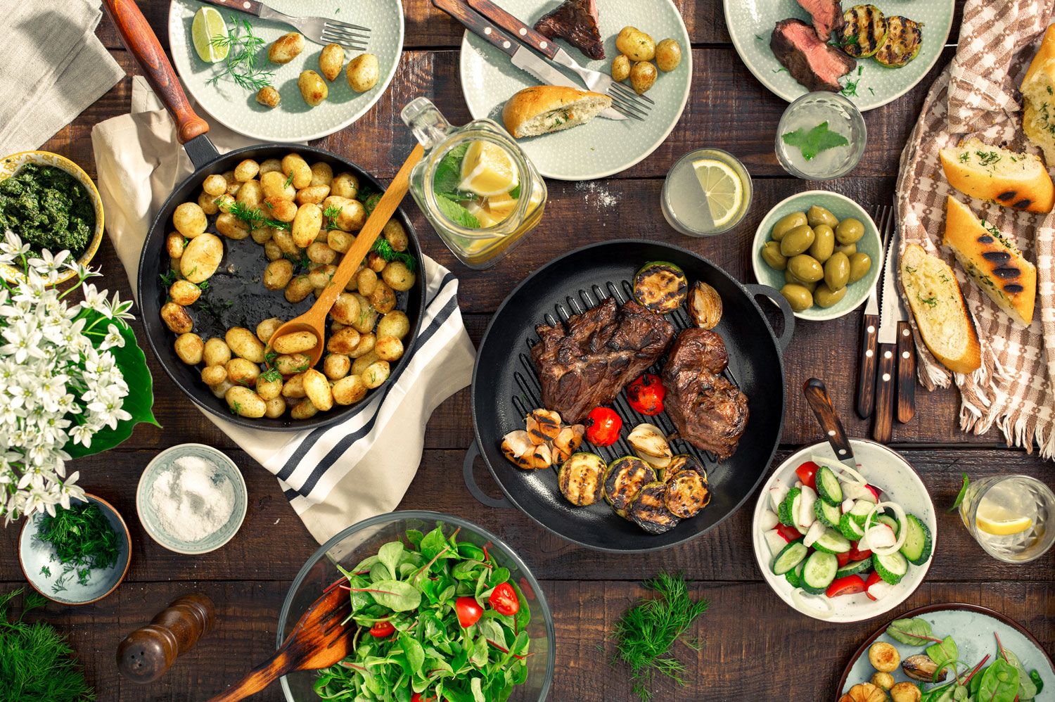 Dinner table with a spread of different foods such as grilled steak, potatoes, salad and bread sitting on a brown table.